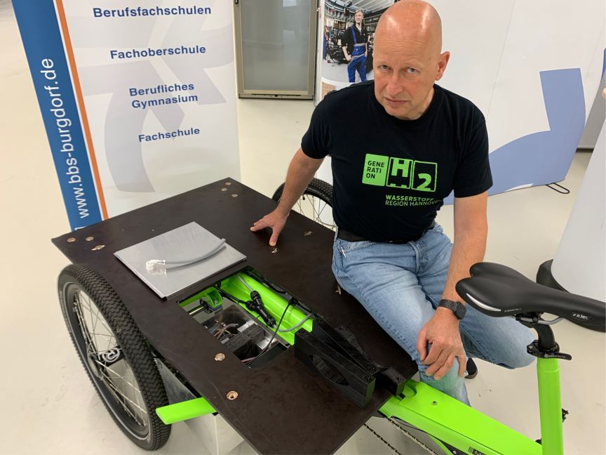 Mann sitzt auf grünem Lastendreirad mit geöffnetem Technikfach, trägt schwarzes H2-Shirt, Hintergrund zeigt Schulbanner.