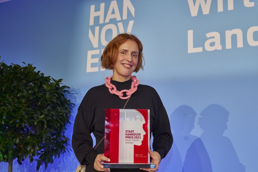 Frau mit roter Halskette hält Auszeichnung „Stadt Hannover Preis 2023" vor blauem Hintergrund mit Hannover-Logo.
