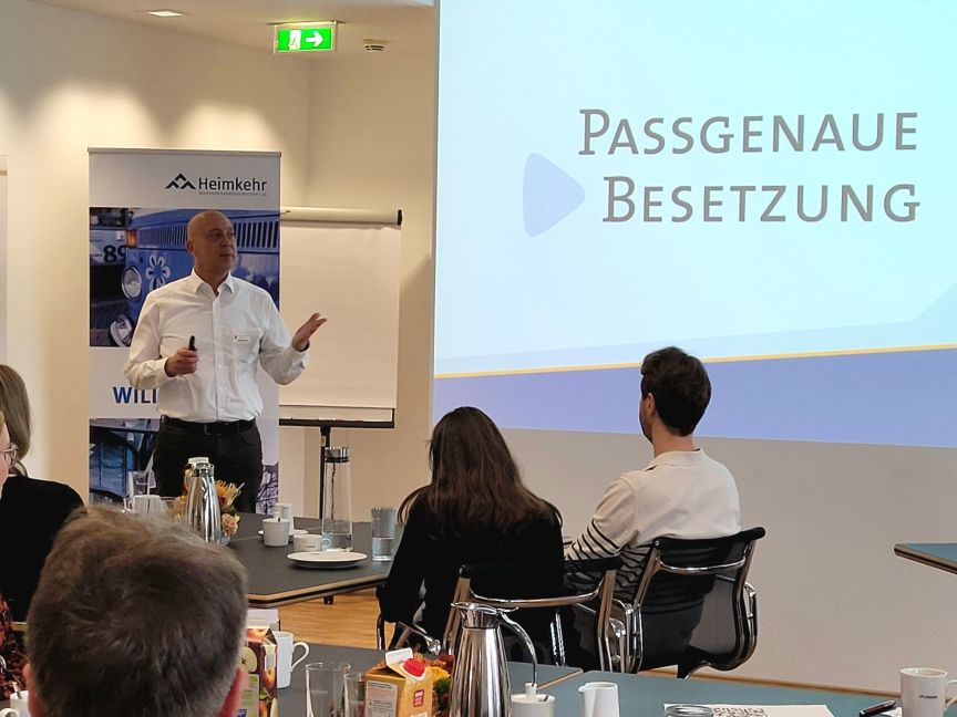 Mann präsentiert vor Publikum. Auf Leinwand: „Passgenaue Besetzung". Im Hintergrund Roll-up mit Logo „Heimkehr".