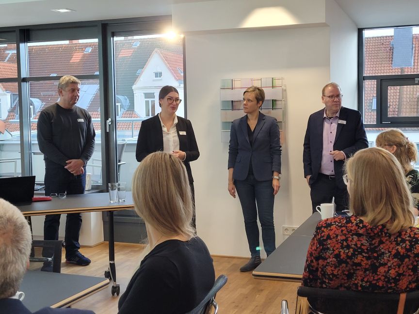 Vier Personen stehen vor einer Gruppe Zuhörender in einem hellen Besprechungsraum mit Fenstern.