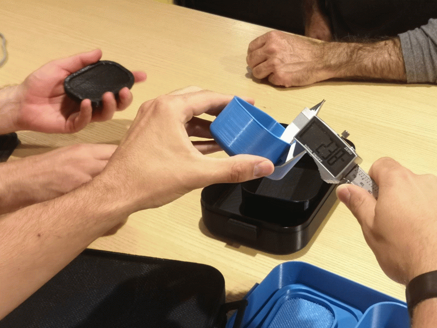 Zwei Personen messen ein blaues 3D-gedrucktes Bauteil mit einem digitalen Messschieber auf einem Tisch.