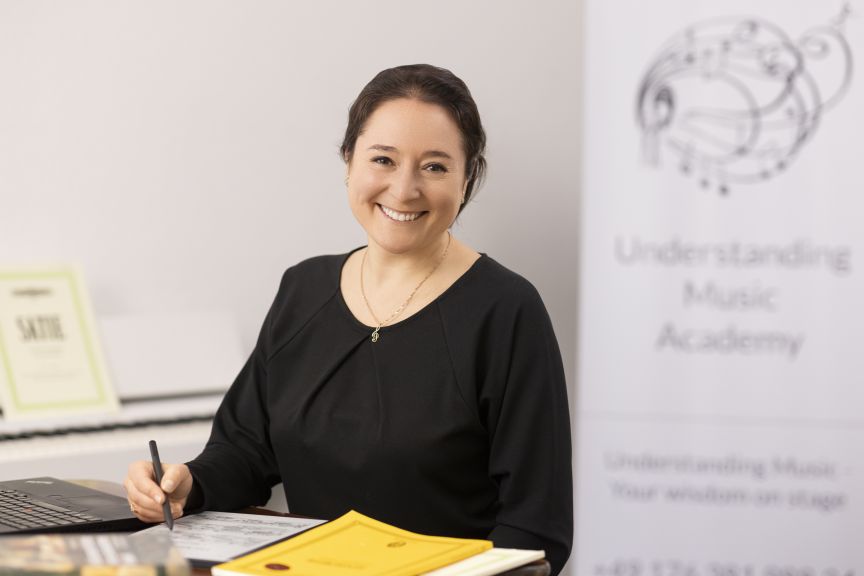 Frau in Schwarz sitzt lächelnd am Schreibtisch mit Stift und Noten; Banner „Understanding Music Academy" im Hintergrund.
