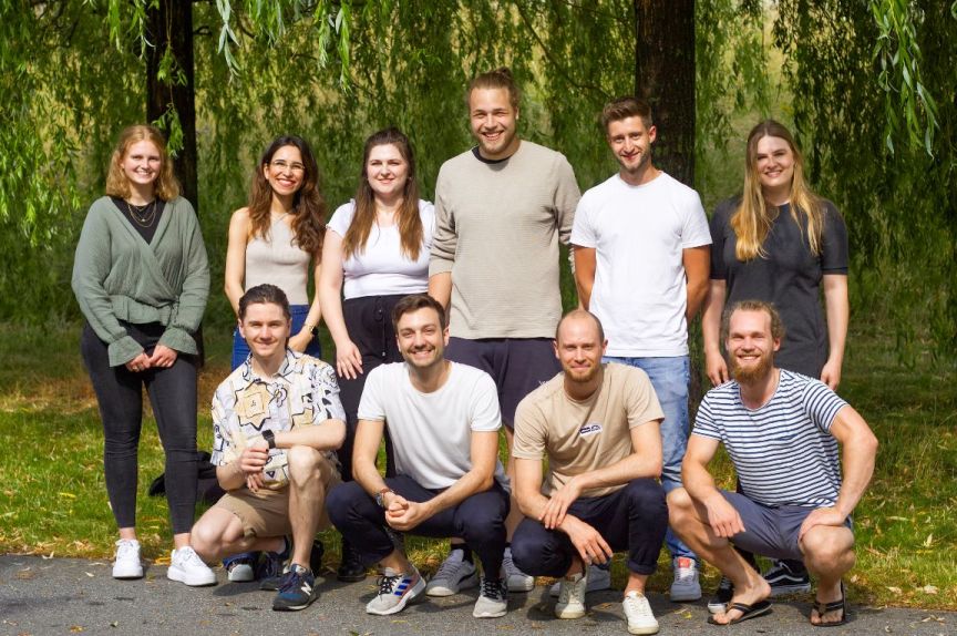 Gruppenfoto von 10 jungen Erwachsenen, 4 kniend vorne, 6 stehend hinten, vor einem Weidenbaum.