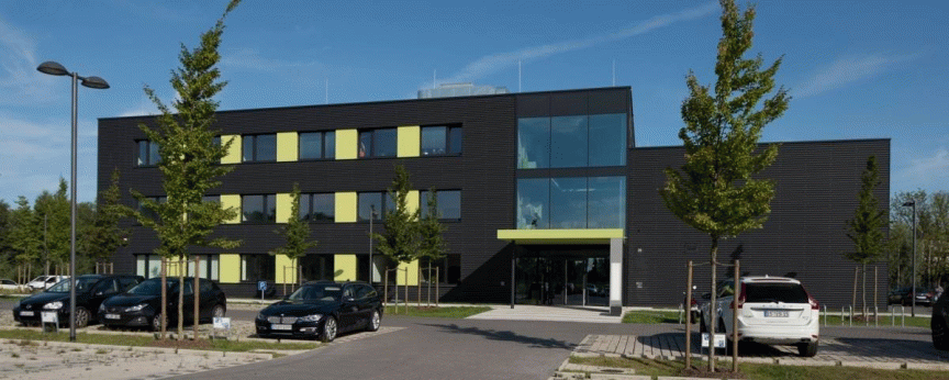 Dreigeschossiges Bürogebäude mit dunkler Fassade und gelben Akzenten, davor Parkplatz mit Autos und Bäumen.
