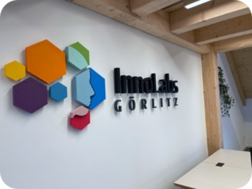 Weißes Firmenlogo „InnoLabs Görlitz" mit bunten Sechsecken an einer Wand in modernem Innenraum mit Holzbalken.